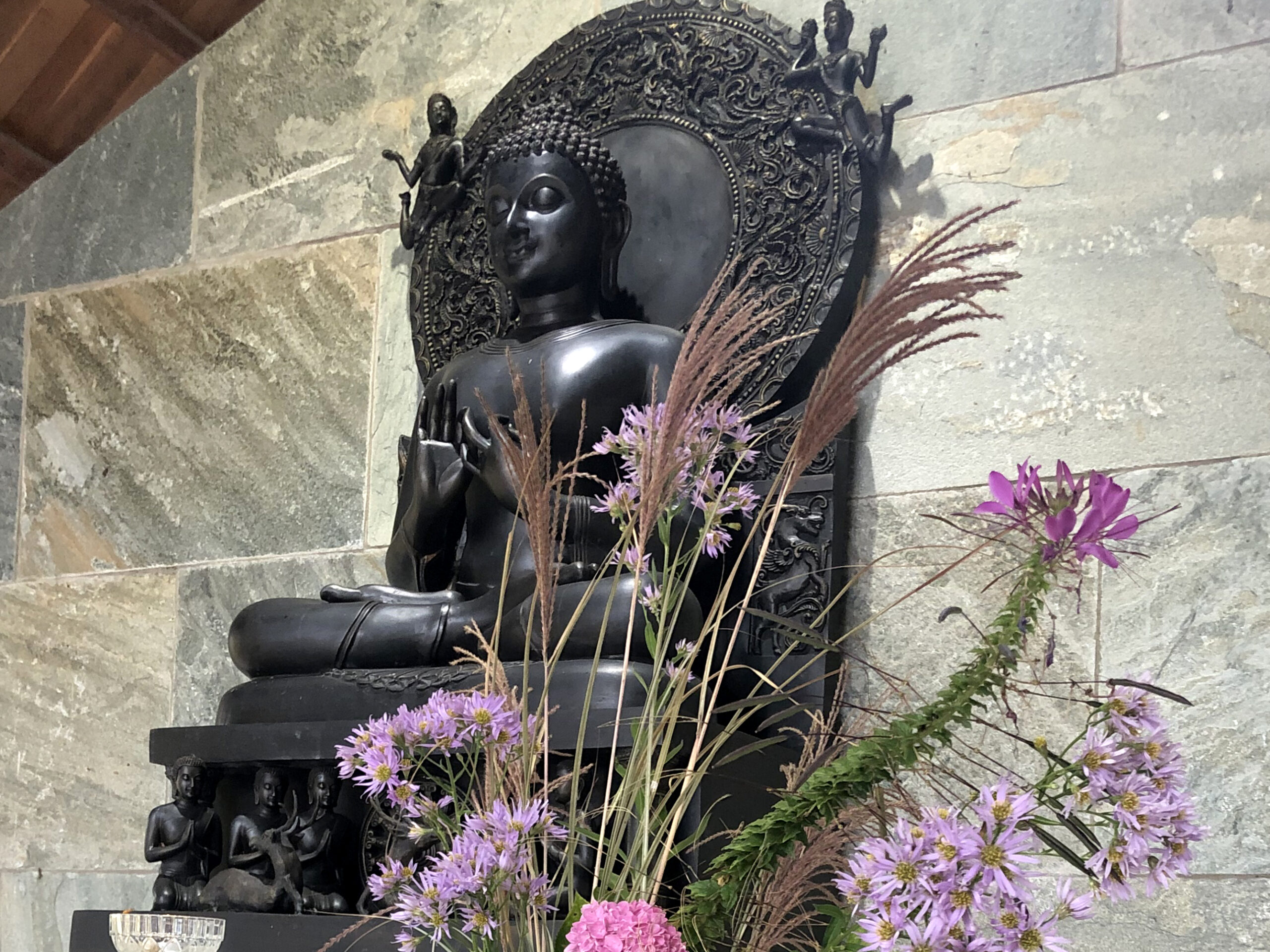 bronze buddha statue against a stone backdrop with purple flowers in front of it